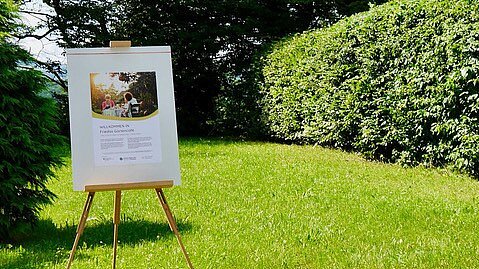 Foto: Im Garten steht eine Flipchart. Darauf ist ein Begrüßungsplakat für Friedas Gartencafé 