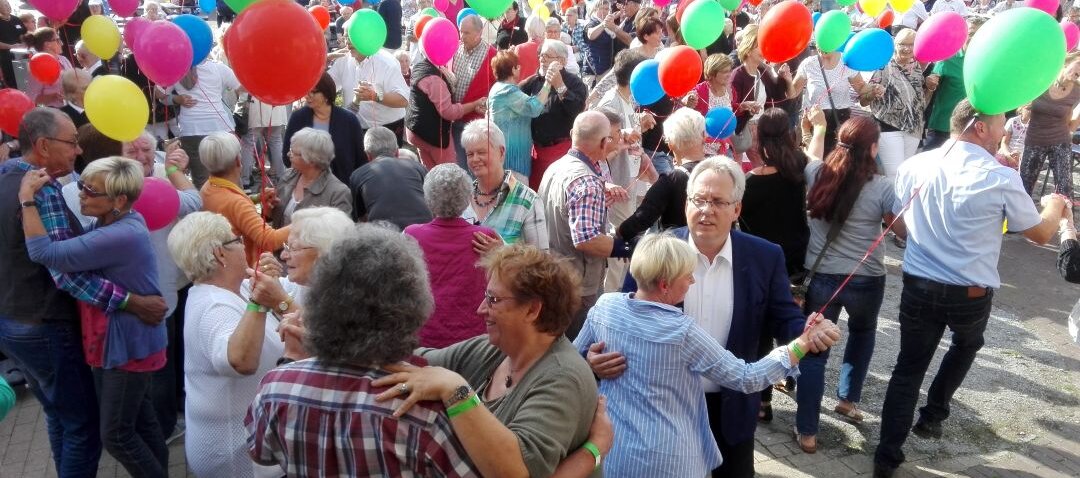 Auf diesem Bild sehen Sie einen Platz, auf dem viele ältere Menschen miteinander tanzen. Viele von ihnen haben einen Luftballon in der Hand.