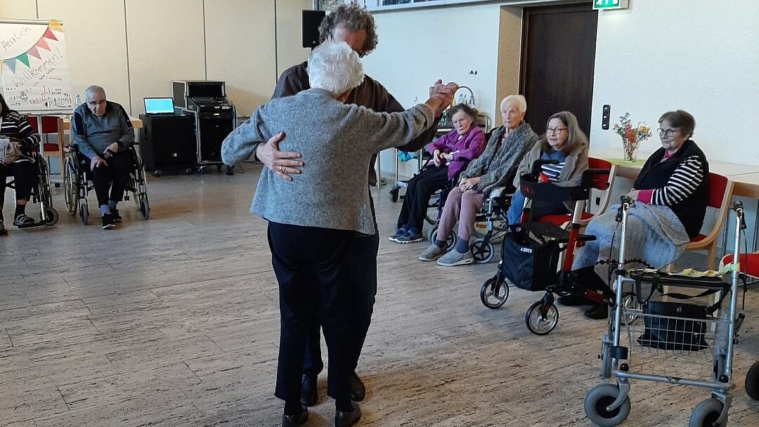 Foto: Mann tanzt mit Seniorin in einem großen Raum, drum herum sitzen Senior*innen und schauen zu.