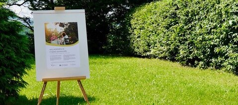 Foto: Im Garten steht eine Flipchart. Darauf ist ein Begrüßungsplakat für Friedas Gartencafé