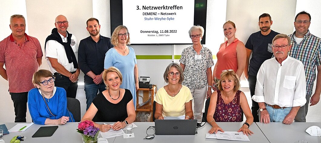 Foto: 4 Frauen sitzen an einem langen Tisch nebeneinander, dahinter stehen 9 Personen. Im Hintergrund ein Bildschirm mit einer Präsentation.