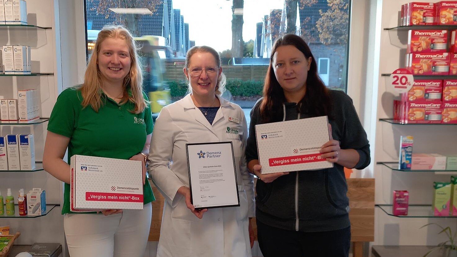 Foto: 3 Frauen stehen in einer Apotheke vor einem Fenster und halten ein Schulungszertifikat und zwei Kartons mit der Aufschrift "Vergiss mein nicht-Box" in die Kamera.
