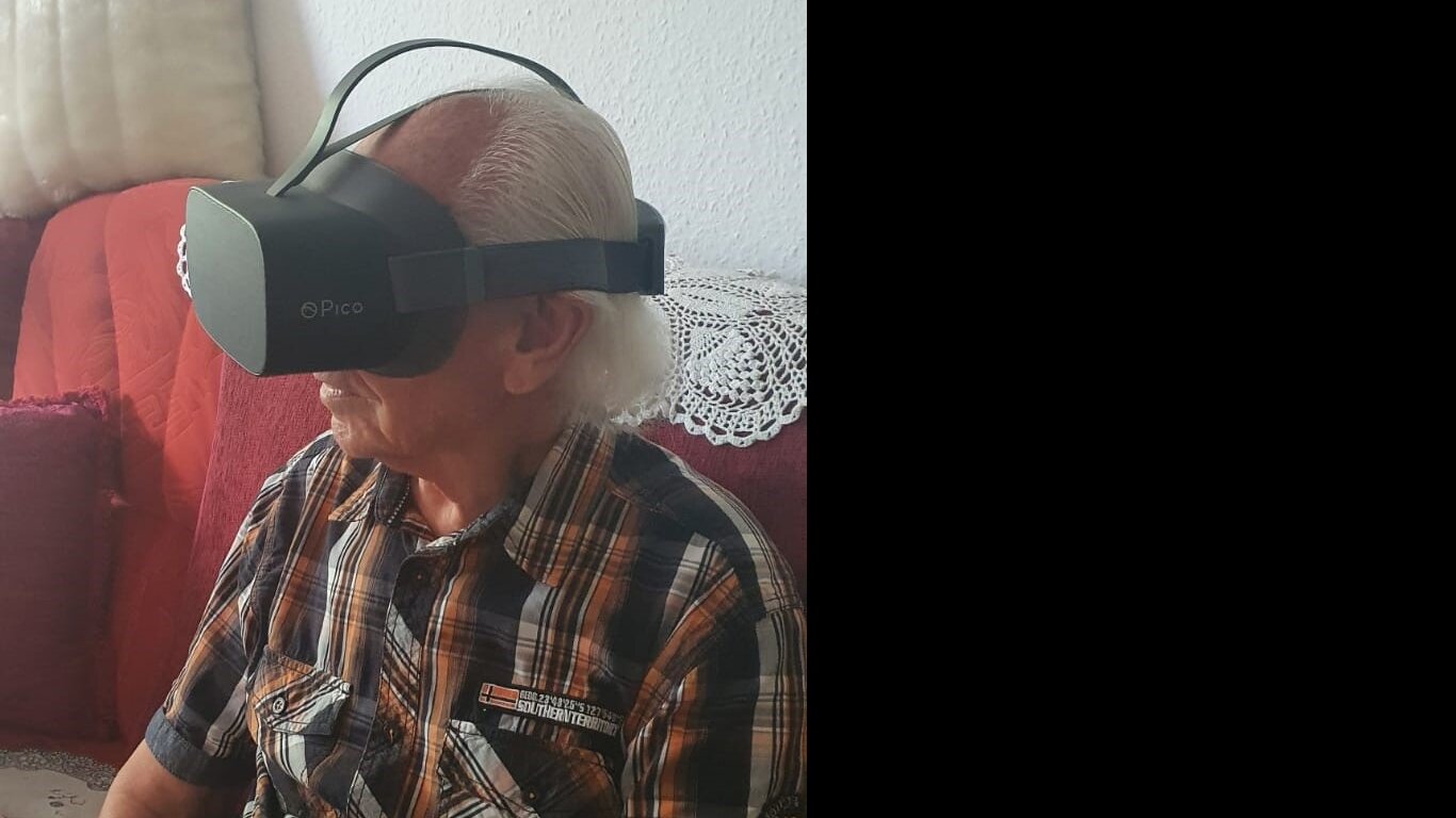 Foto: Ein älterer Mann sitzt auf dem Sofa. Er trägt eine Virtual-Reality-Brille 