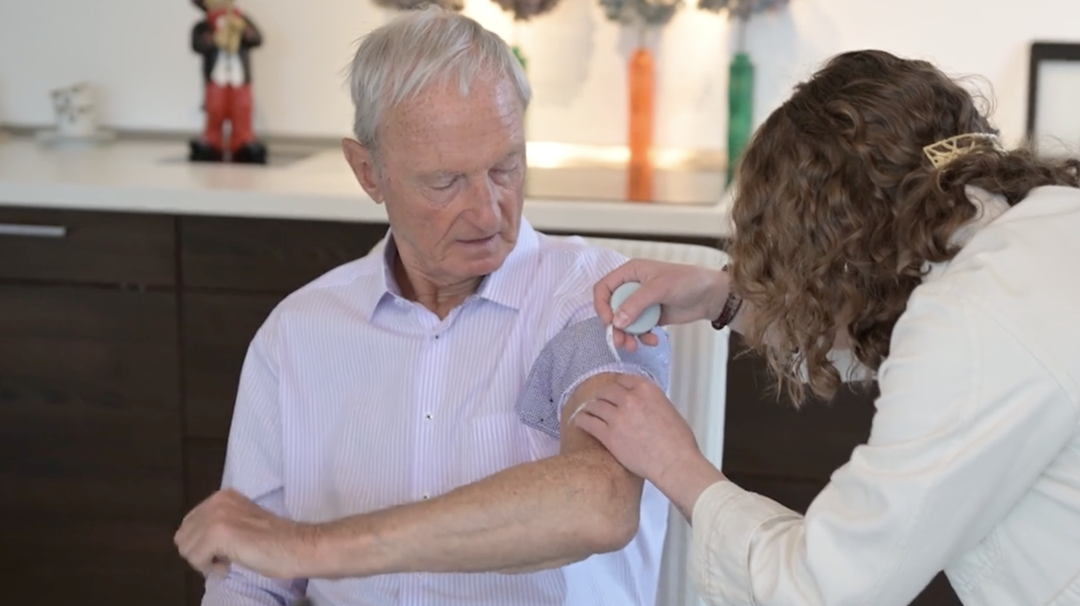 Foto: Senior sitz auf einem Stuhl und wird von Frau in weißem Kittel am Oberarm untersucht.