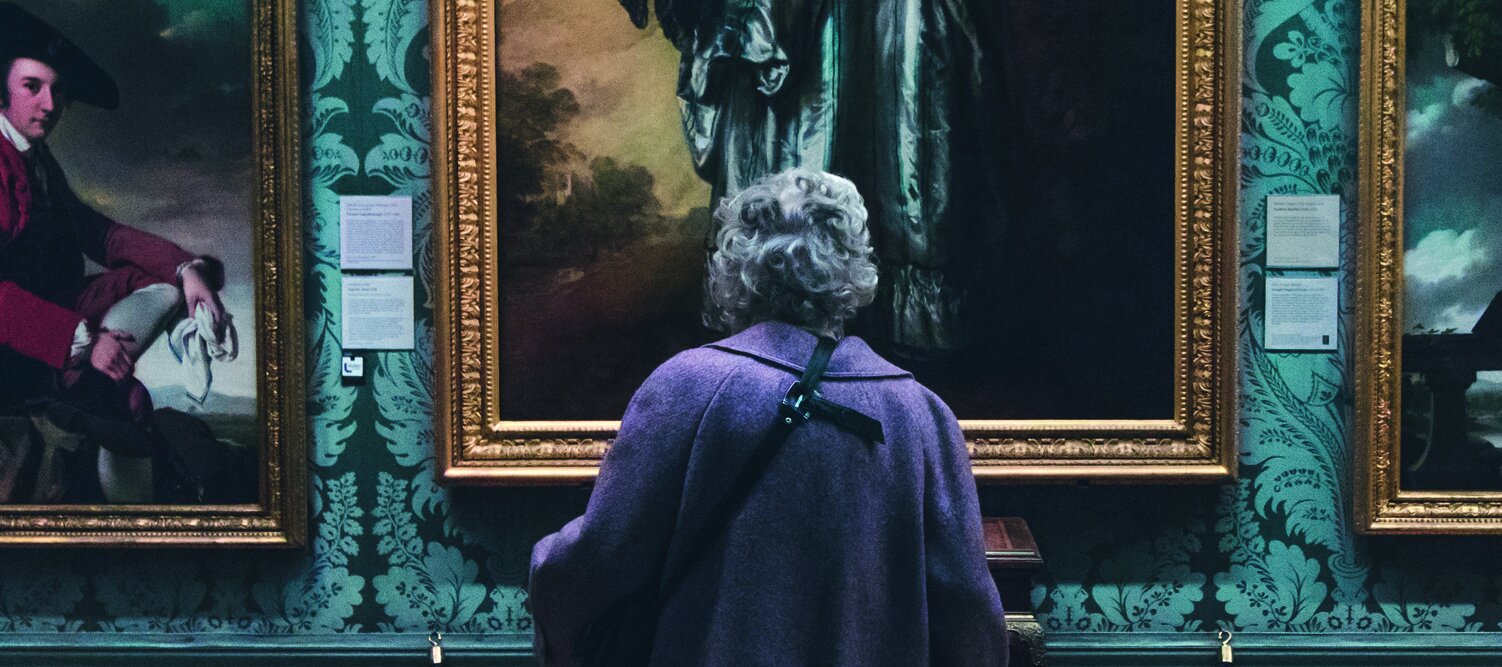 Rückenansicht einer älteren Dame, die im Museum vor einer Wand mit Ölgemälden steht 