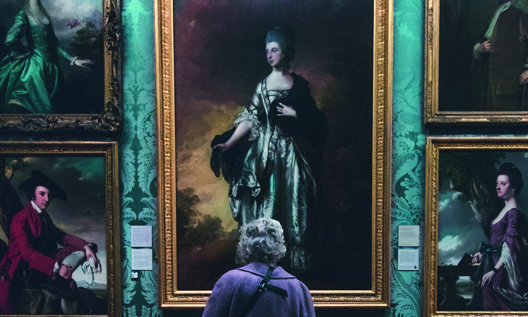 Rückenansicht einer älteren Dame, die im Museum vor einer Wand mit Ölgemälden steht 