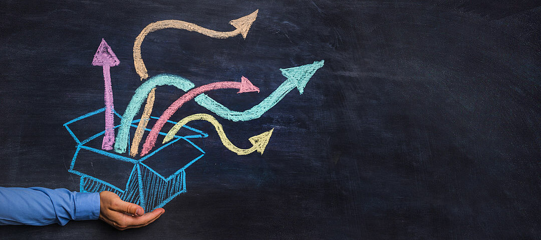 © istock.com_warchi Schwarze Schultafel: ein mit bunter Kreide gemalter geöffneter Karton, aus dem bunte Pfeile herauskommen. Zu sehen ist weiterhin die Hand einer realen Person, die scheinbar den Karton in den Händen hält