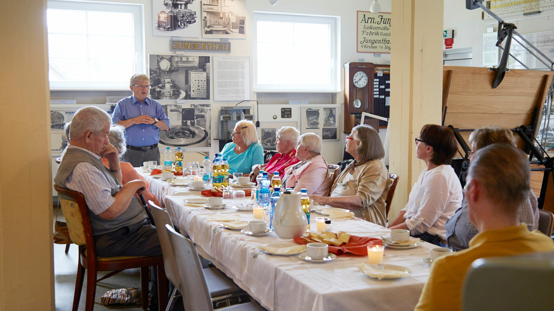 Auf dem Foto sitzen mehrere Senioren an einem langen Tisch. Ein Herr steht 