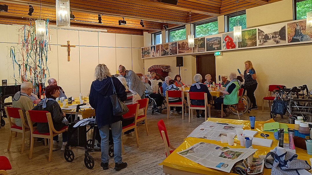 Foto: Senior:innen sitzen an mehreren Tischen mit Getränken auf roten Stühlen, im Hintergrund ein Kruzifix an der Wand