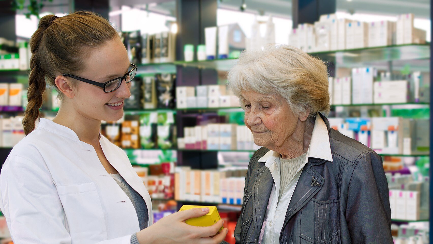 { Foto: Eine junge Apothekerin welche einer älteren Dame ein Medikament zeigt
