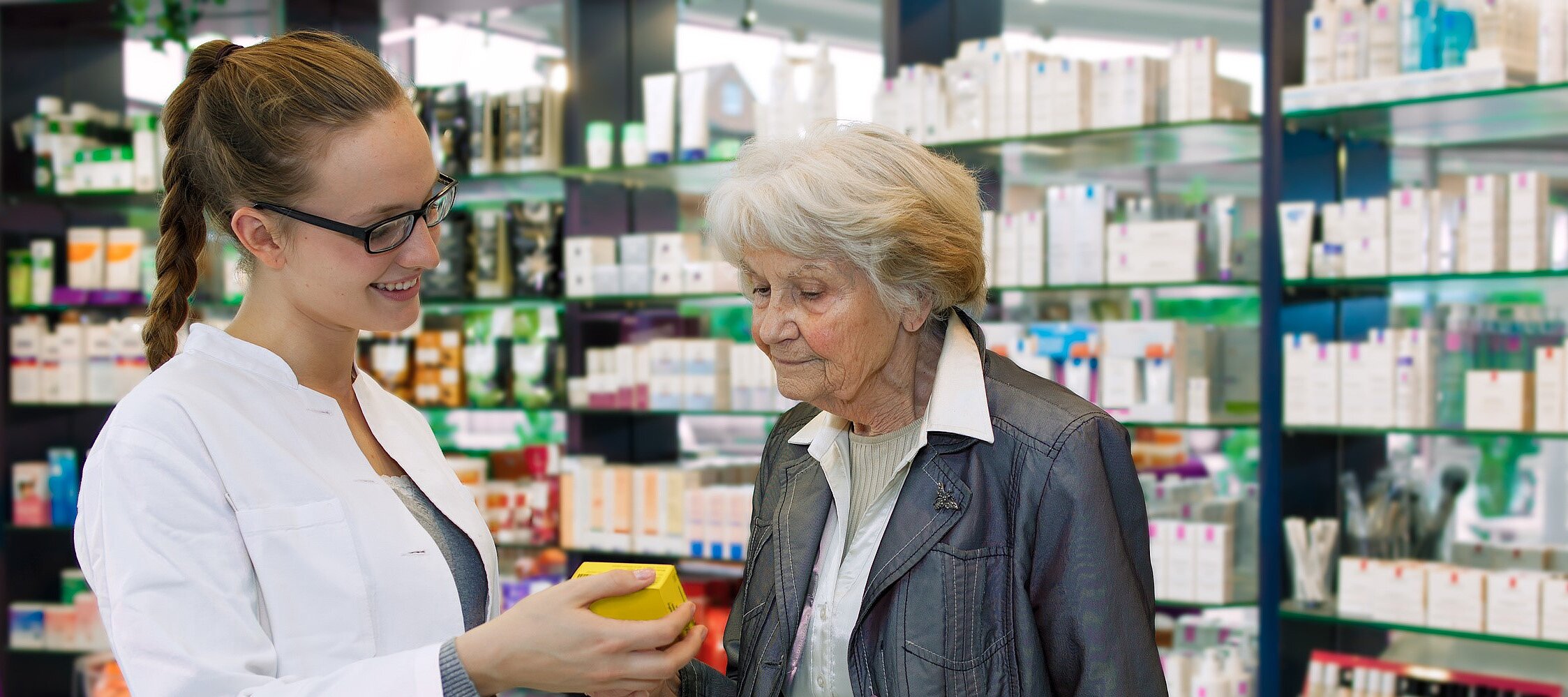 Foto: Eine junge Apothekerin welche einer älteren Dame ein Medikament zeigt