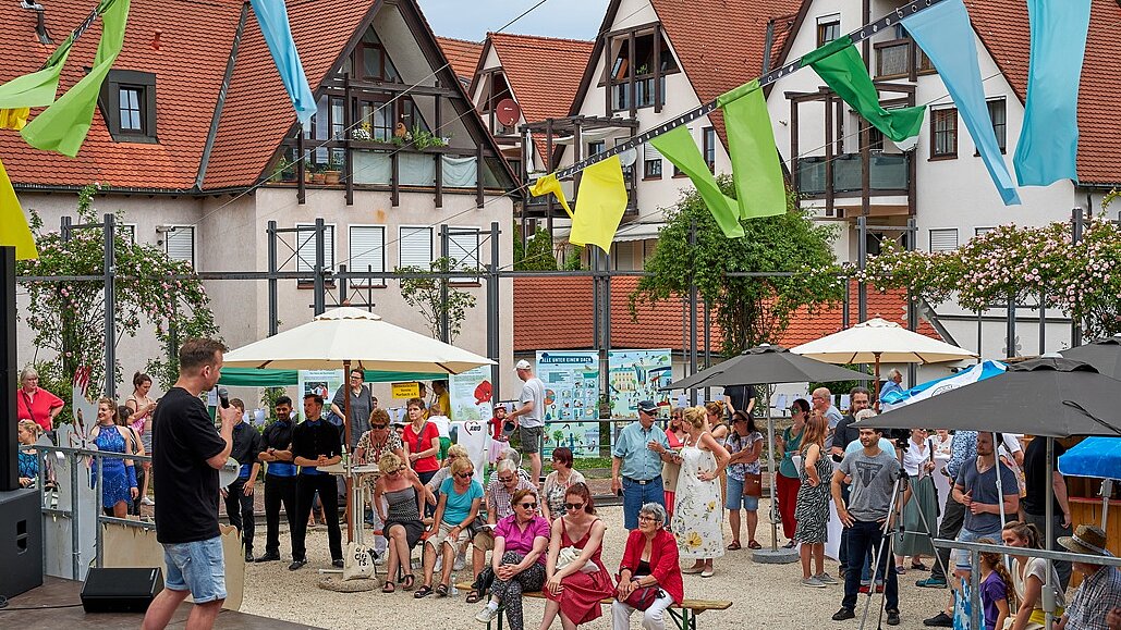 Foto: Menschen schauen auf eine Bühne links am Bildrand auf einem Platz unter Sonnenschirmen vor mehreren Häusern.