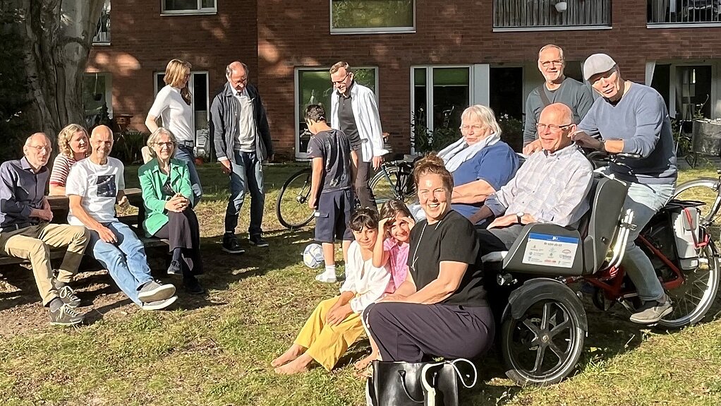 © Susanne Holert-Retzlaff Foto: Auf einem Rasen vor einem Gebäude in der Sonne sitzen vorwiegend Senior*innen, u.a. links auf einer Bank, rechts in einer Fahrradrikscha oder stehen im Hintergrund.