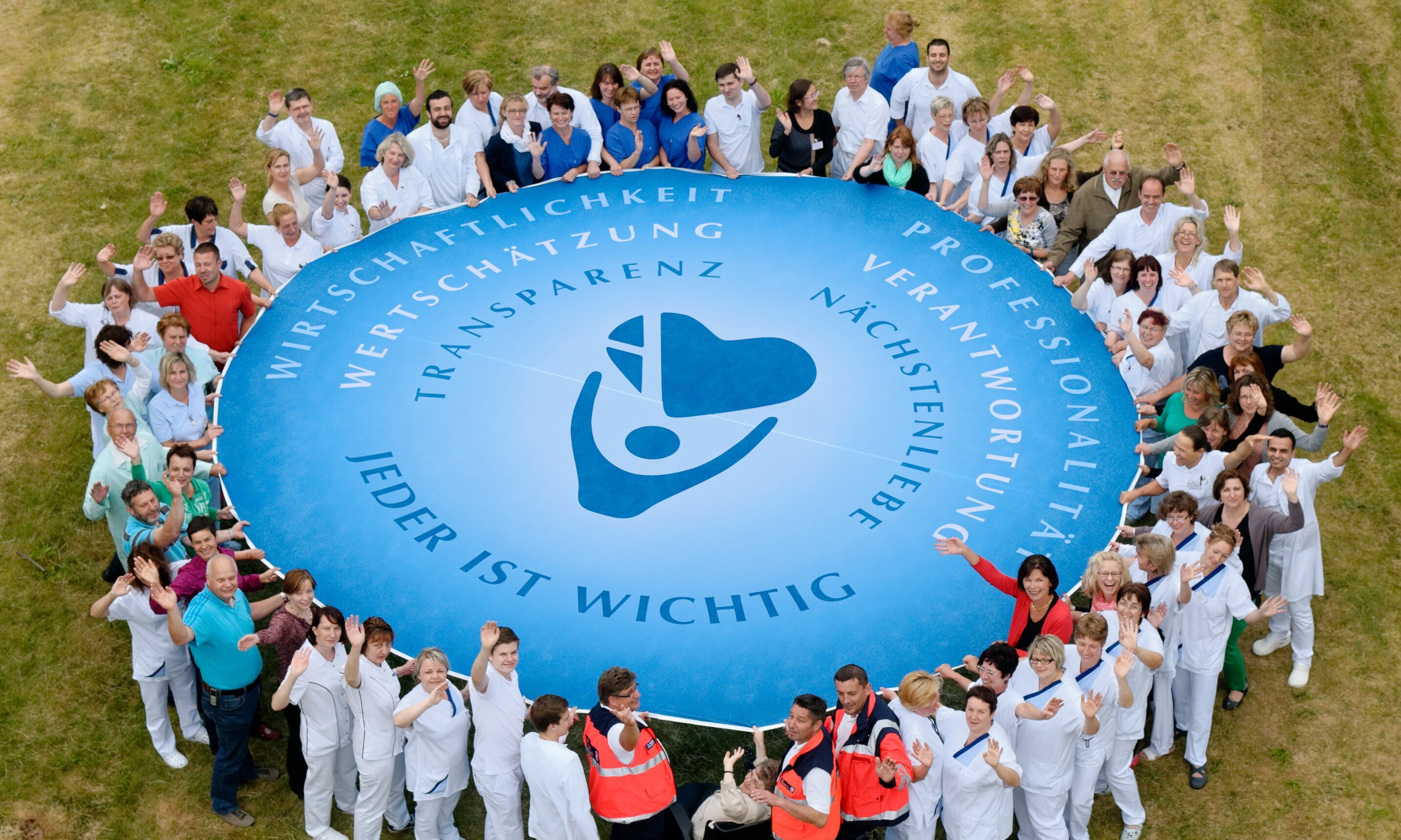 Vogelperspektive: Das Personal des Krankenhauses hält ein Schwungtuch mit Logo der Agaplesion gAG