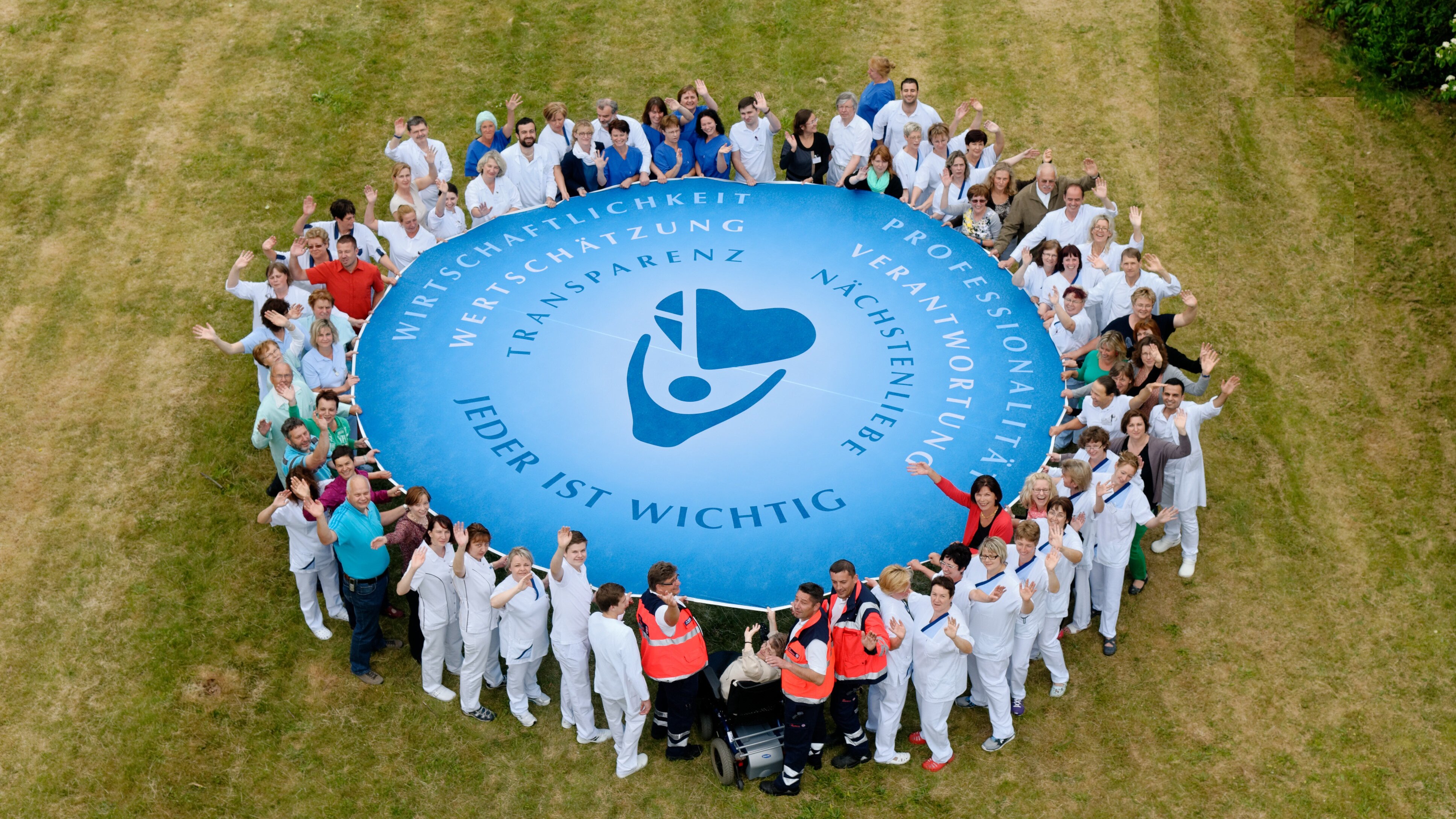 Vogelperspektive: Das Personal des Krankenhauses hält ein Schwungtuch mit Logo der Agaplesion gAG