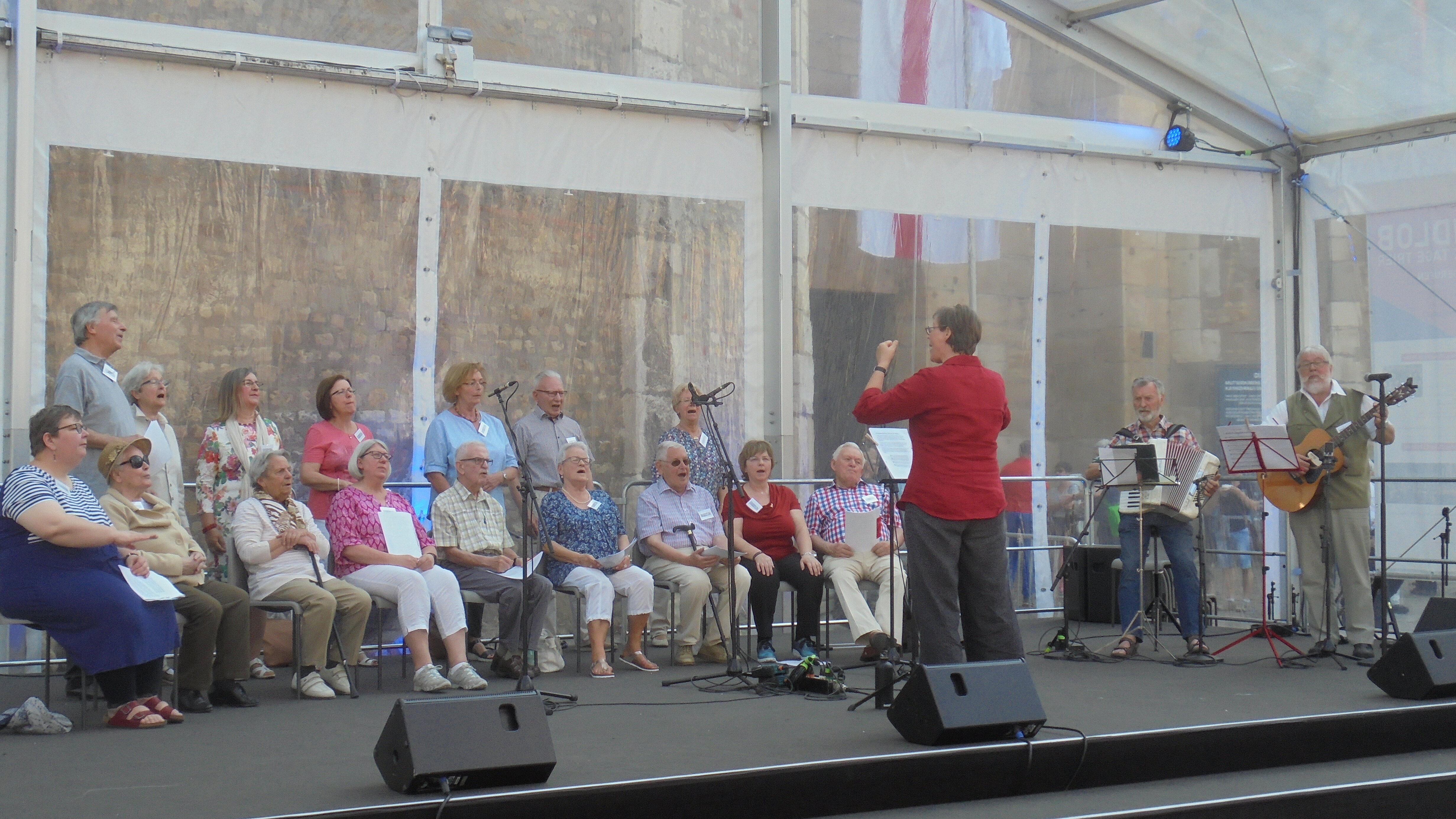 Foto: Foto von einem Chorauftritt mit Dirigentin, einem Akkordeonspieler und einem Gitarristen