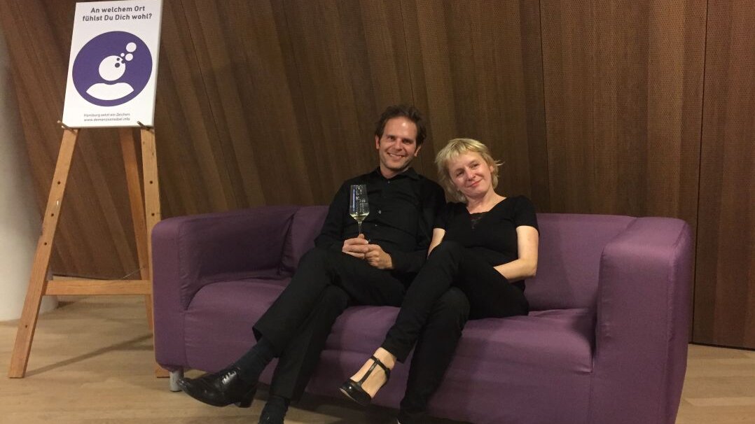 Auf diesem Foto sehen Sie zwei Personen, die auf einem lilafarbenen Sofa sitzen