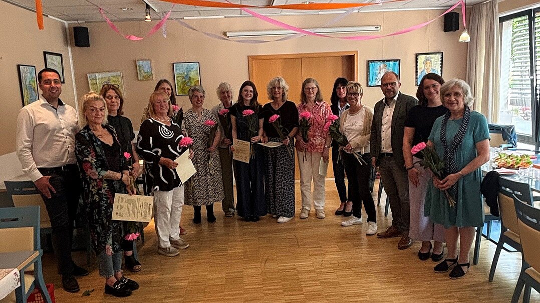 Foto: Eine Gruppe Menschen stehen im Halbkreis mit Urkunden und einer rosa Blume in der Hand.