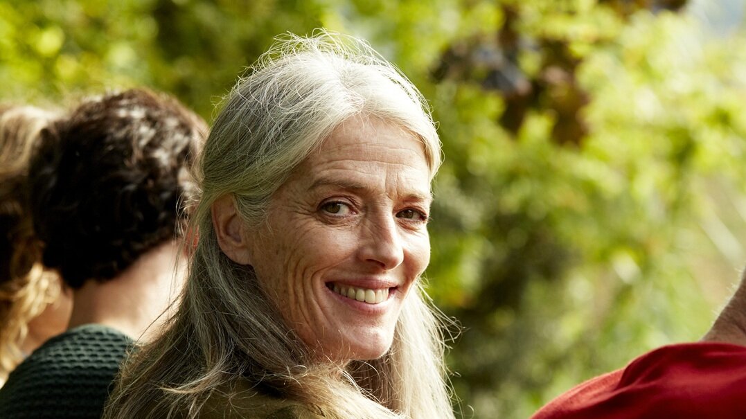 Foto: Frau mit langen grauen Haaren vor grünen Büschen lächelt in Kamera