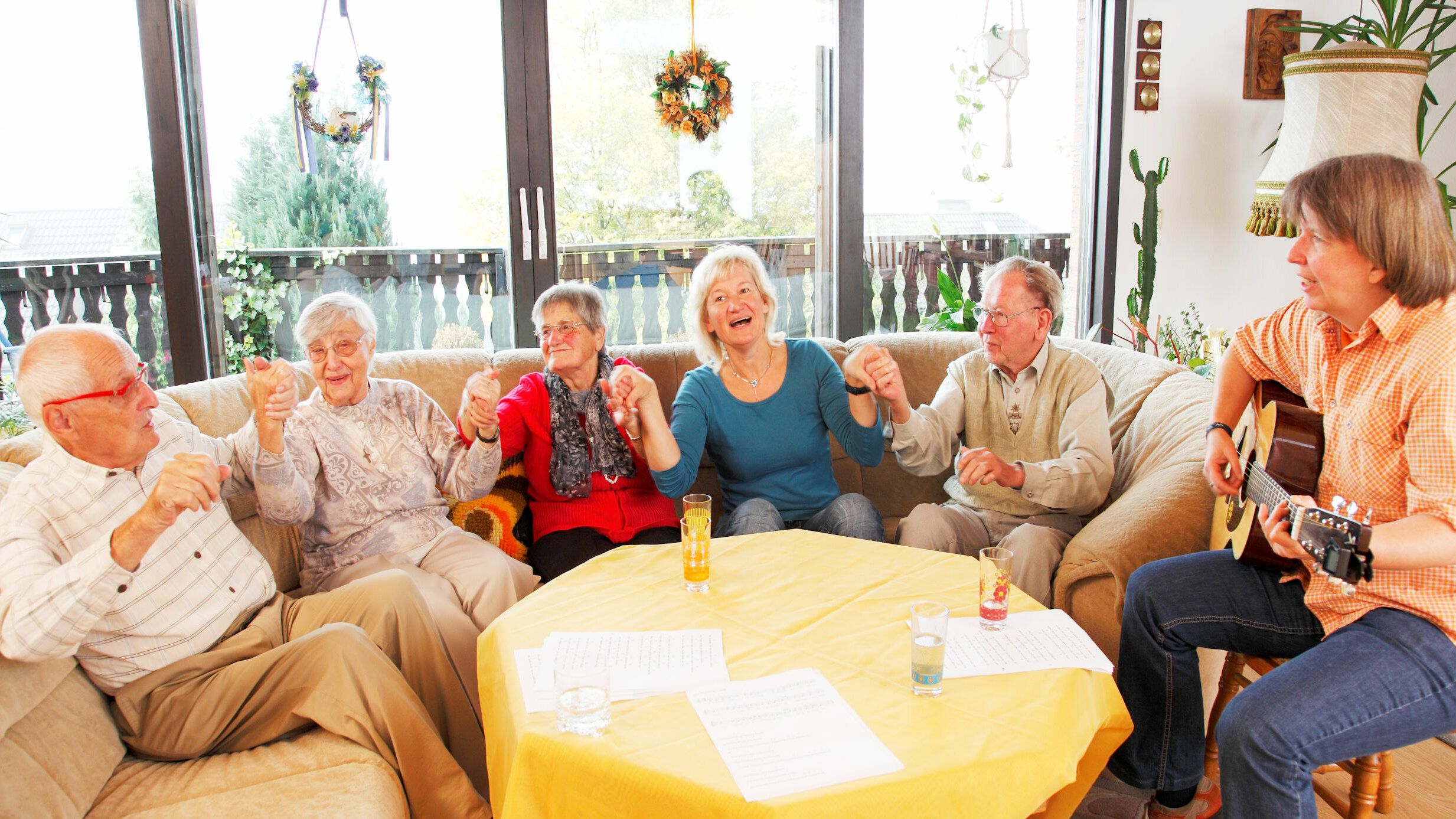 Auf dem Foto sitzen fünf Senioren auf einem Sofa und halten sich an den Händen. Eine weitere Dame sitzt auf einem Stuhl und spielt Gitarre