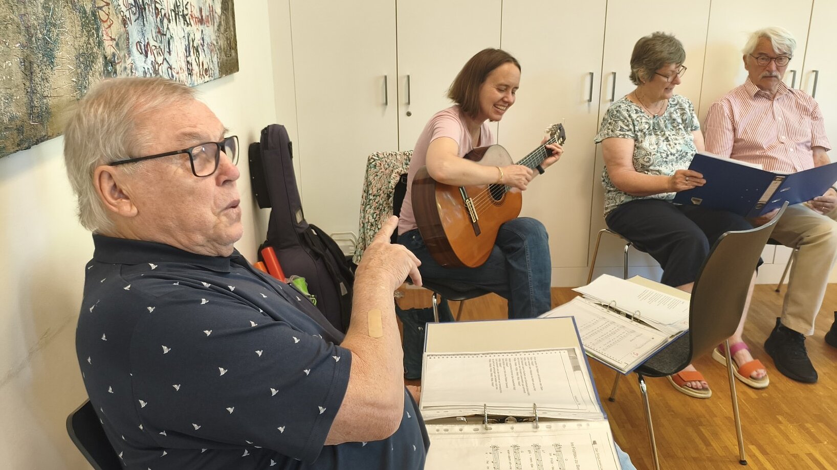 Foto: 3 Senior*innen sitzen in einem Raum neben einer jungen Frau mit Gitarre