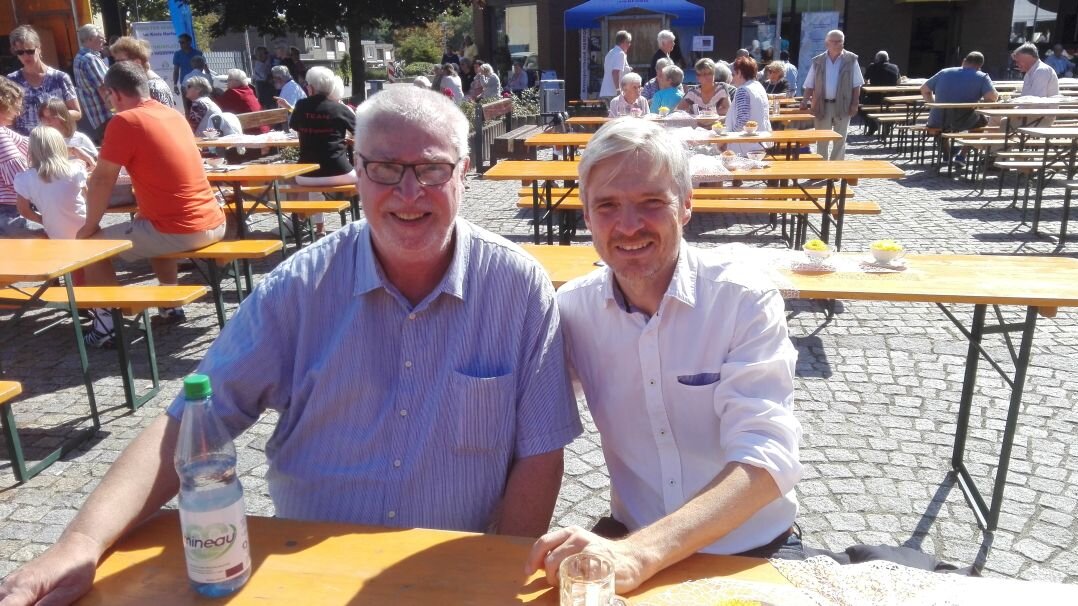 Auf diesem Foto sehen Sie zwei Männer, die an einem Biertisch sitzen. 