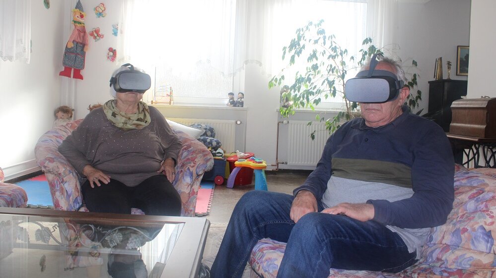 Foto: Eine ältere Frau und ein älterer Mann sitzen in einer Sitzgruppe und tragen Virtual-Reality-Brillen 