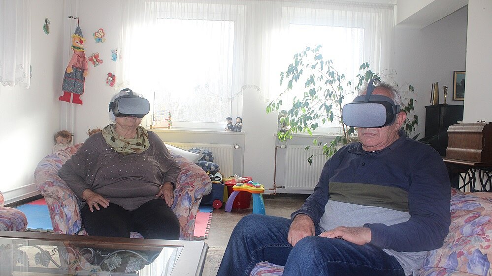 Foto: Eine ältere Frau und ein älterer Mann sitzen in einer Sitzgruppe und tragen Virtual-Reality-Brillen 