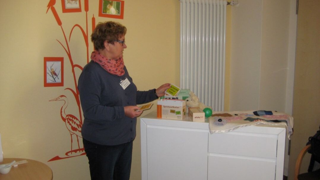 Foto: Demenzbegleiterin in einem gelb gestrichenen Patientenzimmer. Auf einem Rollwagen sind Spiele zum Gedächtnistraining