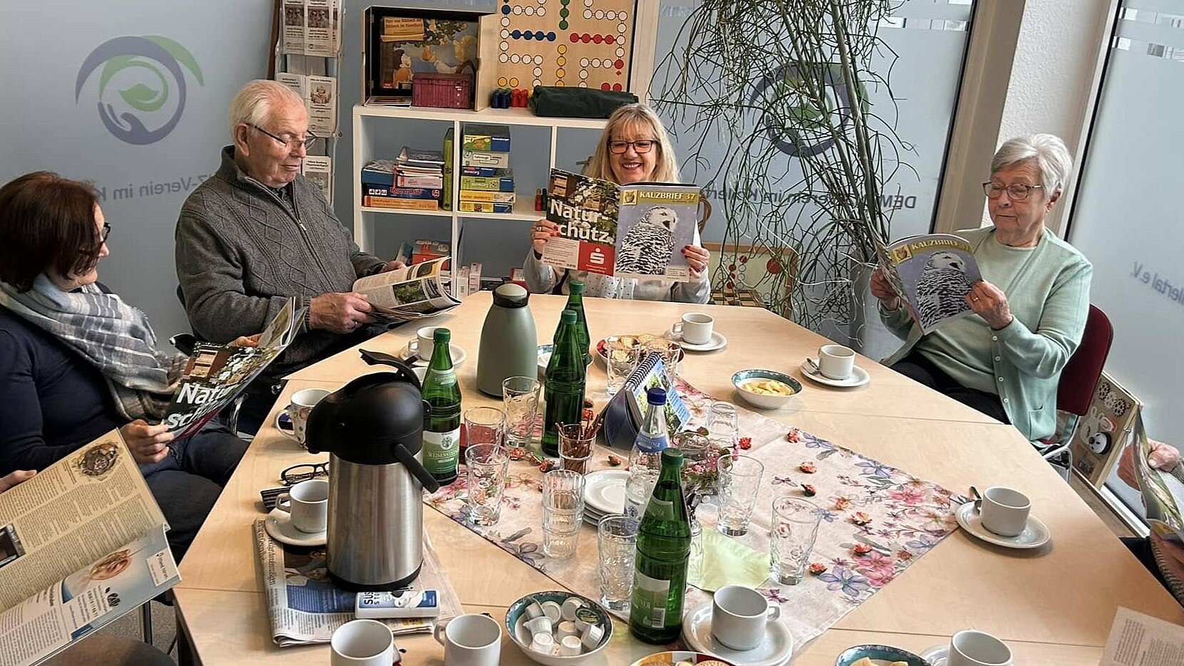 Foto: Mehrere Personen aus unterschiedlichen Generationen sitzen an einem Tisch mit Getränken und lesen in einer Zeitschrift.