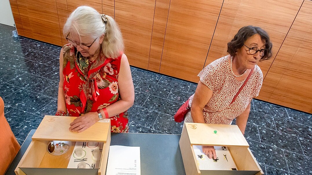 Foto: 2 Seniorinnen sitzen an einem Tisch und tasten in Holzkisten eines Demenzparcours