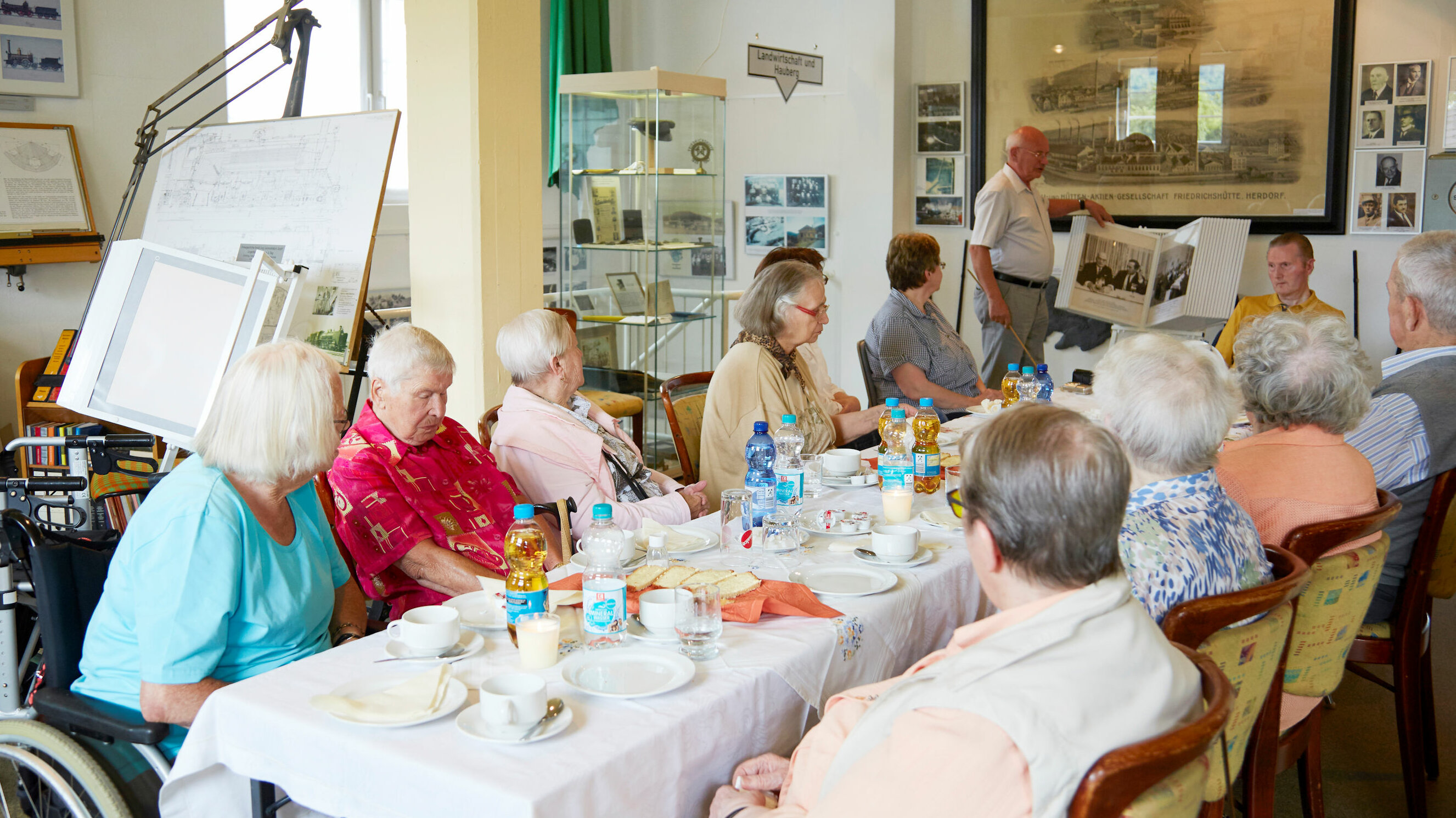 Auf dem Foto hat sich eine Gruppe von Senioren an einem langen Tisch versammelt. Ein weiterer älterer Herr zeigt alte Fotos
