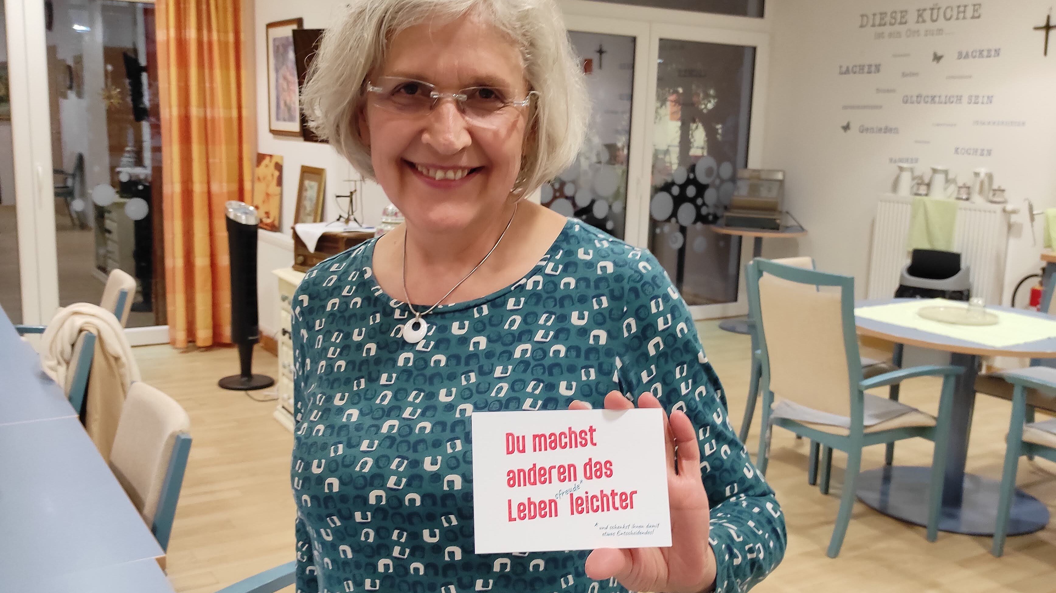 Foto: Seniorin steht in einem Aufenthaltsraum mit Tischen und Stühlen und hält eine Postkarte mit dem Aufdruck "Du machst anderen das Leben leichter" in die Kamera.