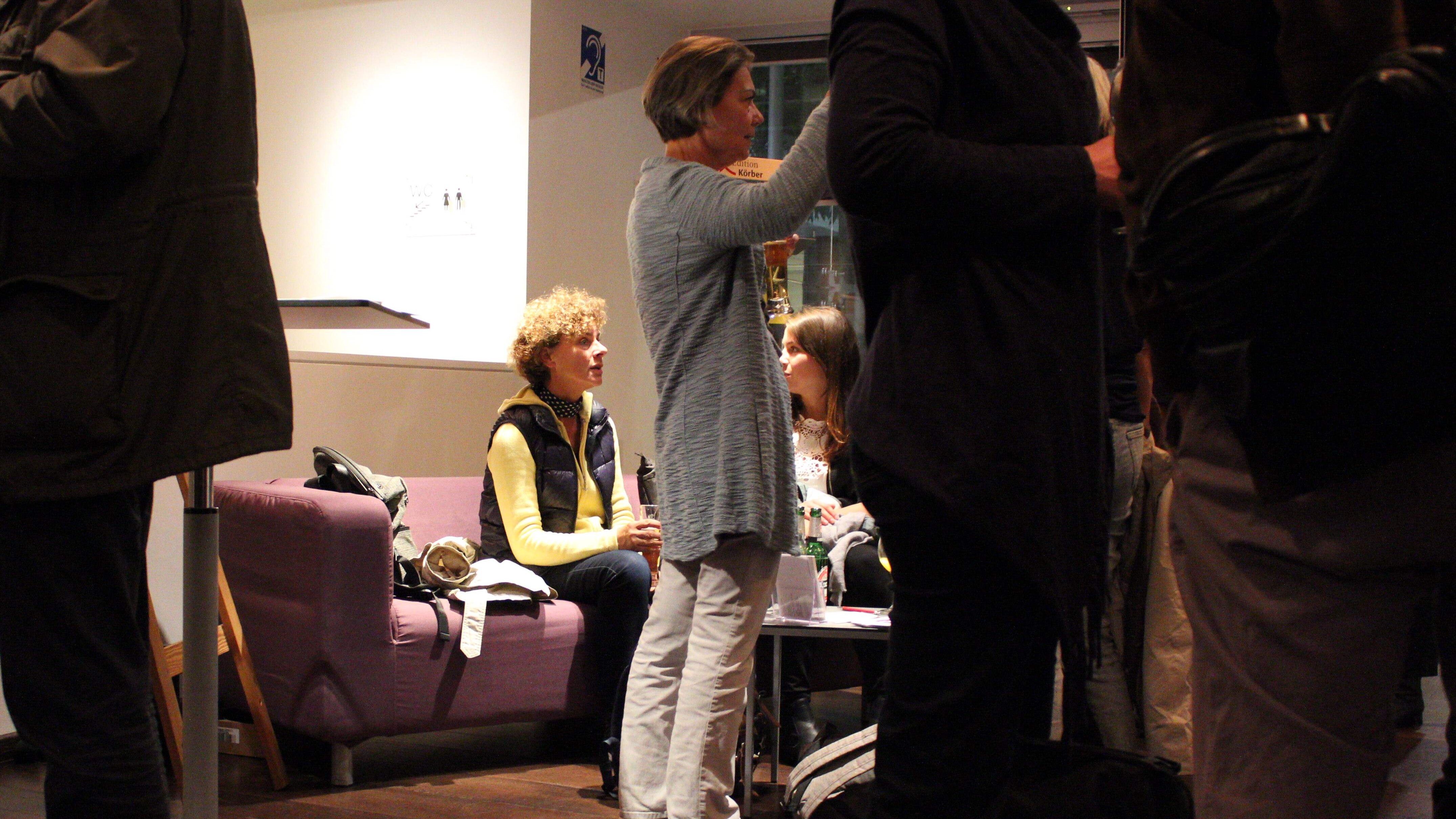 Foto: Im Vordergrund stehen mehrere personen, im Hintergrund sitzen zwei Personen auf einem lila Sofa