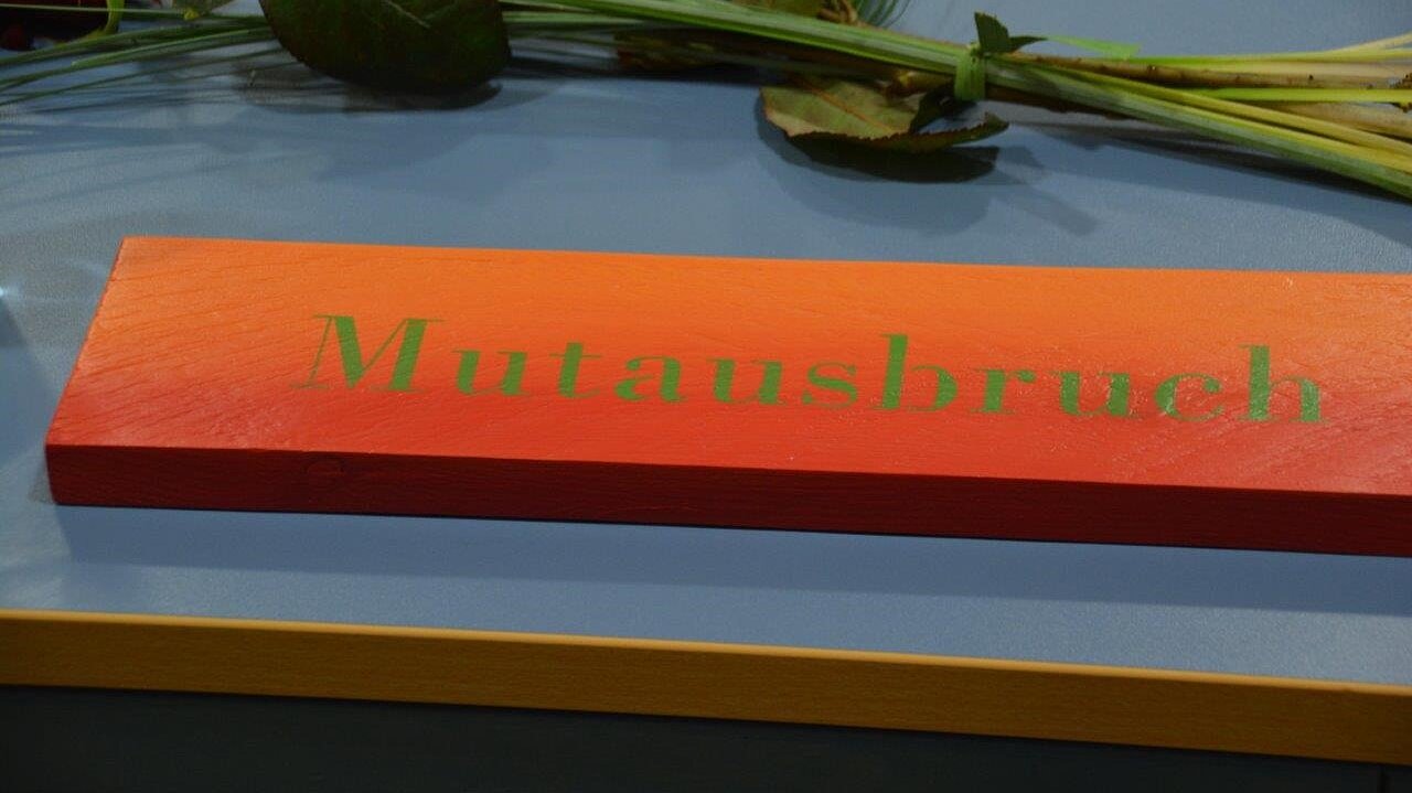 Foto: Holzschild mit grünem Aufdruck "Mutausbruch" liegt auf grauem Tisch mit Blumenschmuck.