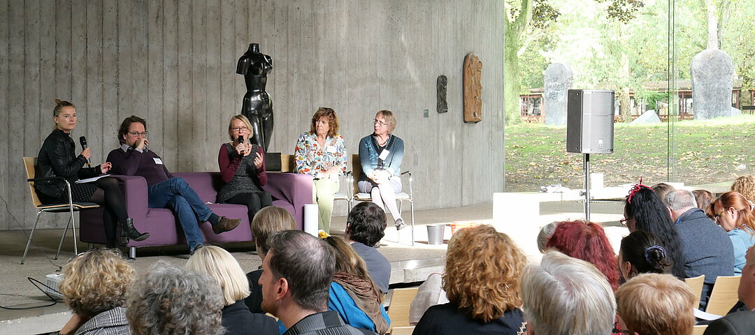 Auf dem Foto sieht man viele Menschen in einem Museumsraum sitzen. Auf der Bühne sitzen Personen auf einem lila Sofa. 