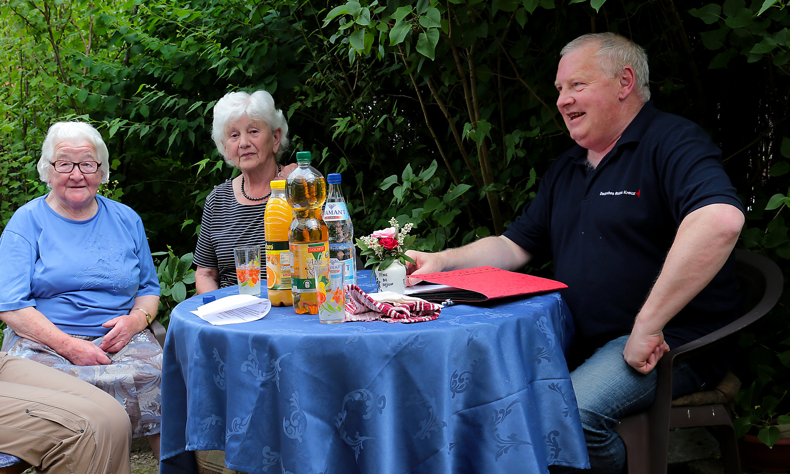 Ein Mitarbeiter des Deutschen Roten Kreuzes sitzt mit zwei älteren Damen an einem Gartentisch 
