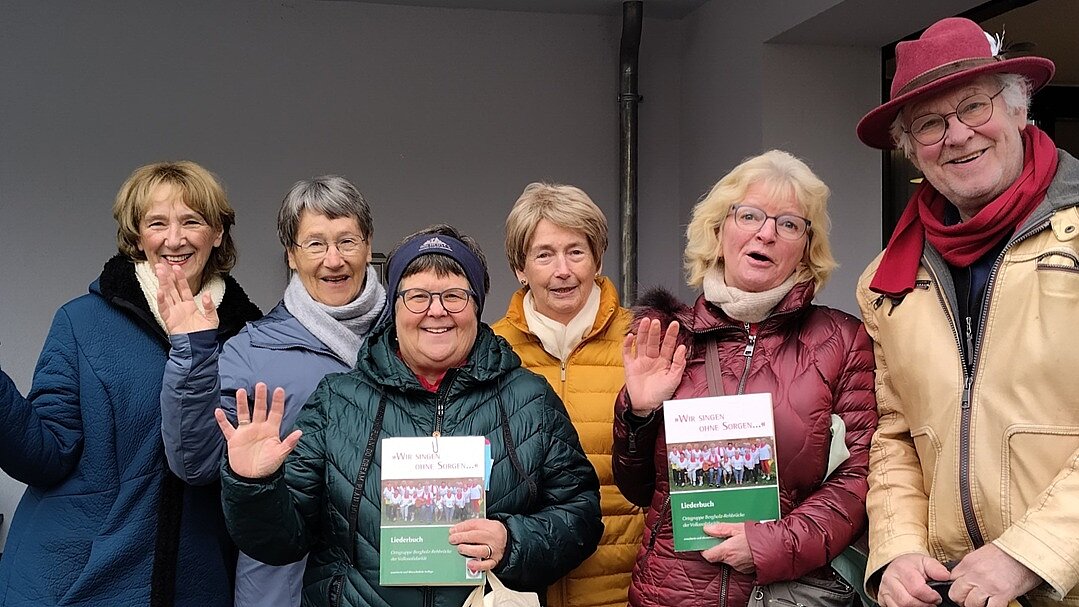 Die Singegruppe der Volkssolidarität erfreut die Bewohnerinnen und Bewohner der Seniorenresidenz mit gemeinsamem Gesang. Foto: 6 Senior*innen winken lächelnd in die Kamera, 2 halten eine Broschüre in der Hand.