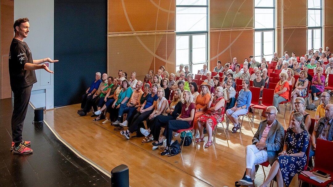 Foto: Mann steht links am Bildrand auf Bühne vor Publikum in großem Saal mit roten Stühlen
