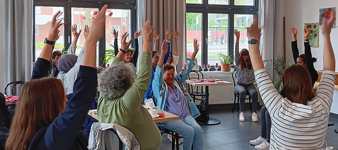 Foto: Senioren sitzen in einem Stuhlkreis in einem Raum und heben die Arme in die Höhe.
