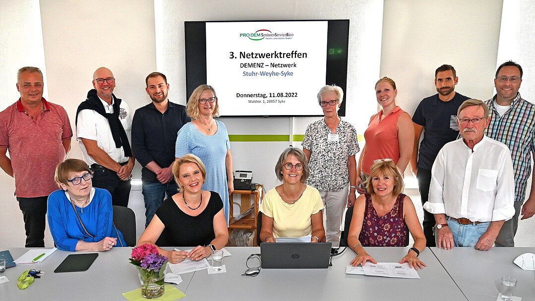 Gemeinde Syke: Drittes Netzwerktreffen im August 2022 Foto: 4 Frauen sitzen an einem langen Tisch nebeneinander, dahinter stehen 9 Personen. Im Hintergrund ein Bildschirm mit einer Präsentation.