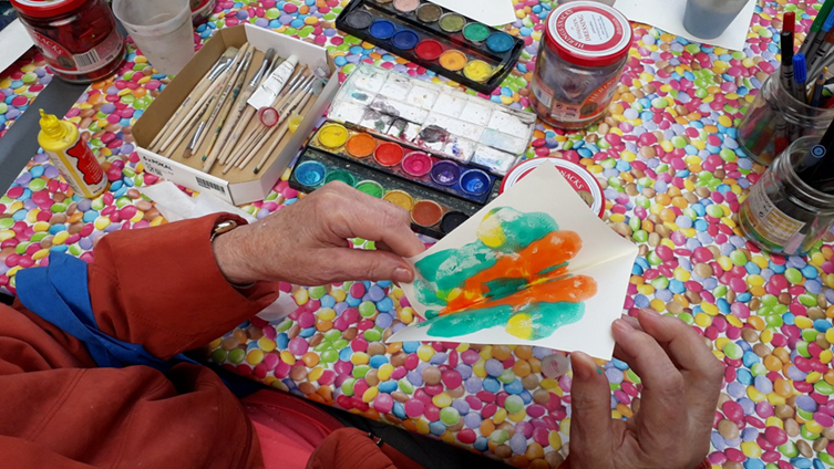 Foto: Tisch mit verschiedenen Malutensilien, die Hände einer älteren Frau halten ein selbstgemaltes Bild 