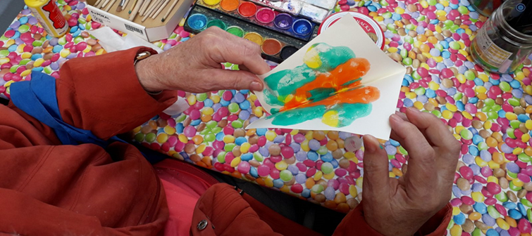 Tisch mit Malutensilien: Zu sehen sind die Hände einer älteren Frau, die ein Wasserfarbenbild in der Hand hält