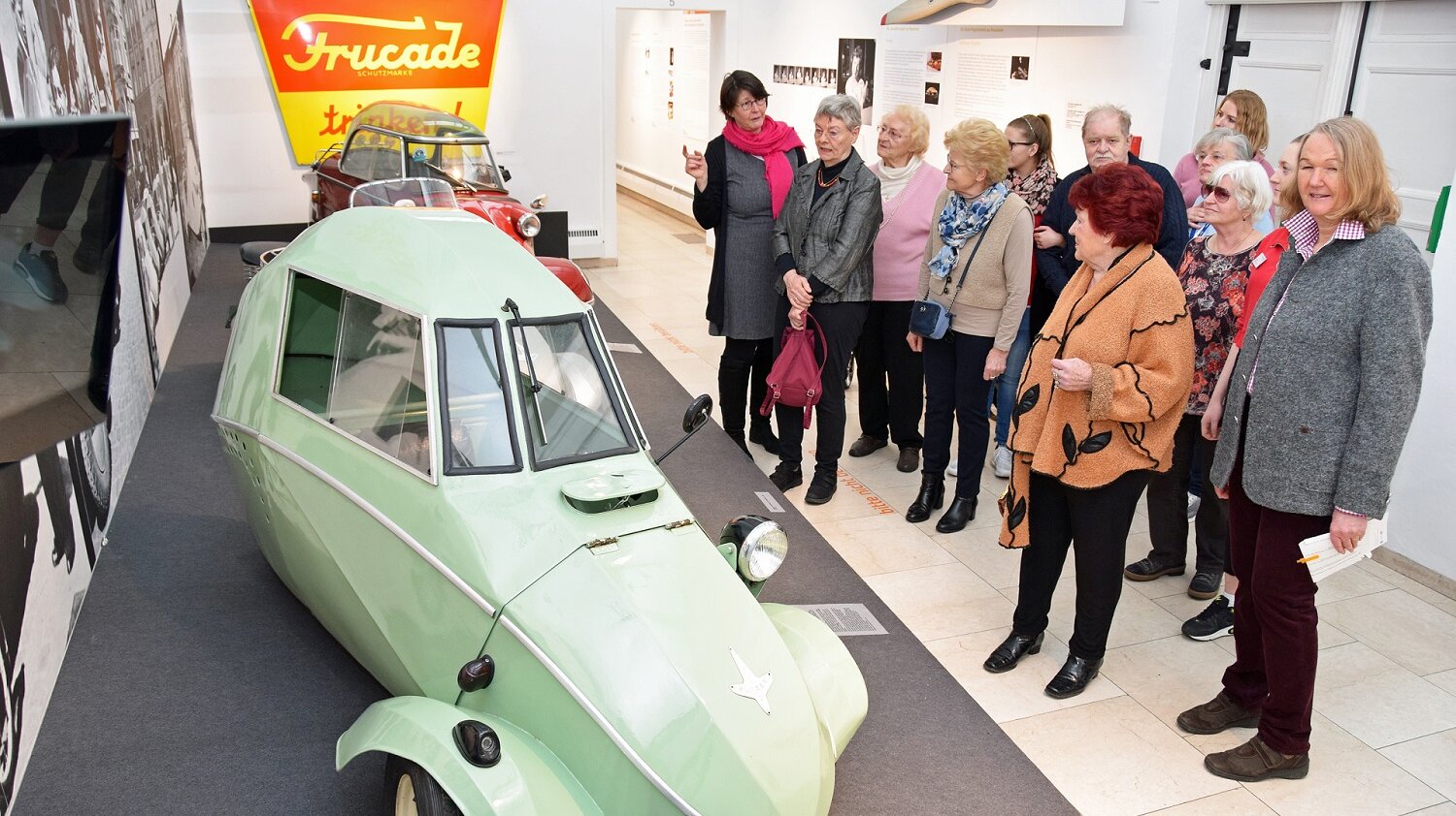 Seniorinnen und Senioren des BRK Heims Rosenheim erkunden zusammen mit der Historikerin Lydia Zellner (im Bild links) eine Ausstellung in der Rosenheimer Galerie zum Thema 100 Jahre Design in Rosenheim. Foto: Eine Gruppe von Seniorinnen und Senioren betrachtet in der Städtischen Galerie Rosenheim einen lindgrünen Oldtimer
