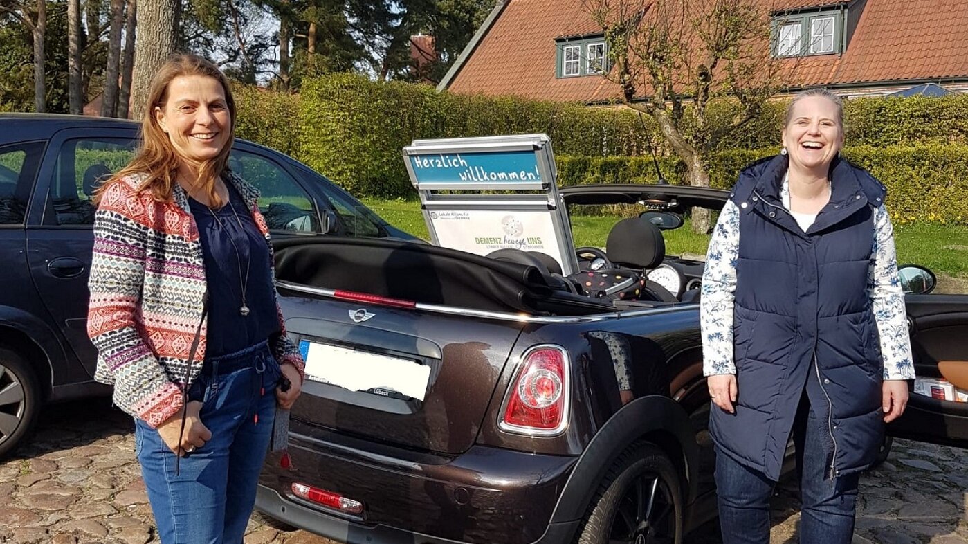 Foto: Parkendes Auto mit offenem Verdeck und Poster der Lokalen Allianz auf dem Rücksitz. Rechts und links stehen zwei Frauen und lachen in die Kamera.