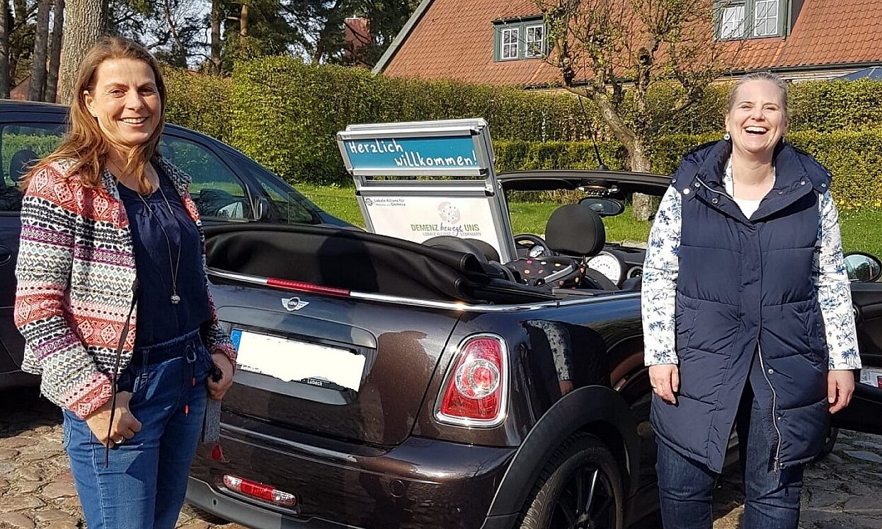 Foto: Parkendes Auto mit offenem Verdeck und Poster der Lokalen Allianz auf dem Rücksitz. Rechts und links stehen zwei Frauen und lachen in die Kamera.