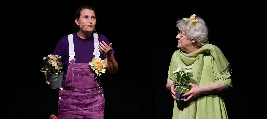 Zwei Schauspielerinnen auf der Bühne. Eine der beiden ist lila gekleidet, die andere grün. Beide haben einen Blumentopf in der Hand