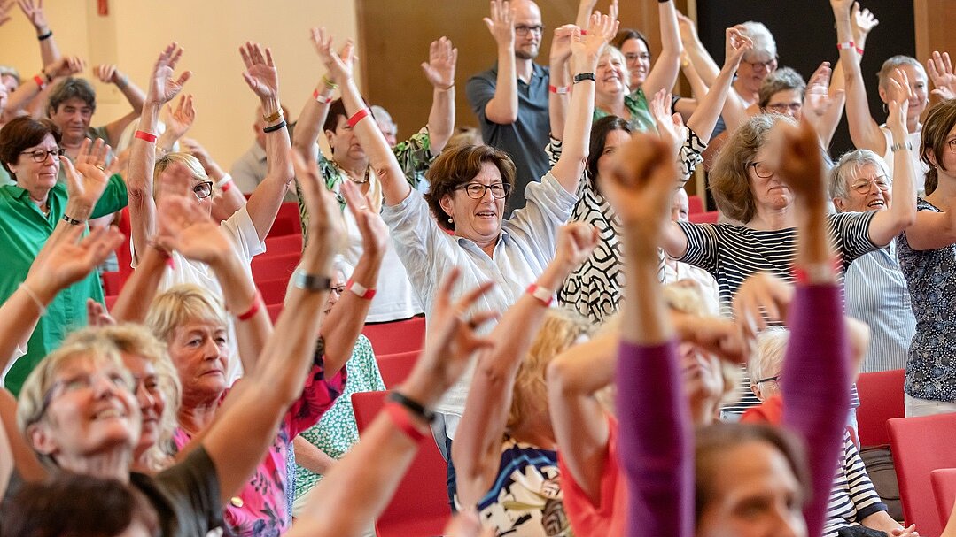 Foto: Menschen sitzen auf oder stehen vor roten Stühlen und haben beide Arme hochgestreckt