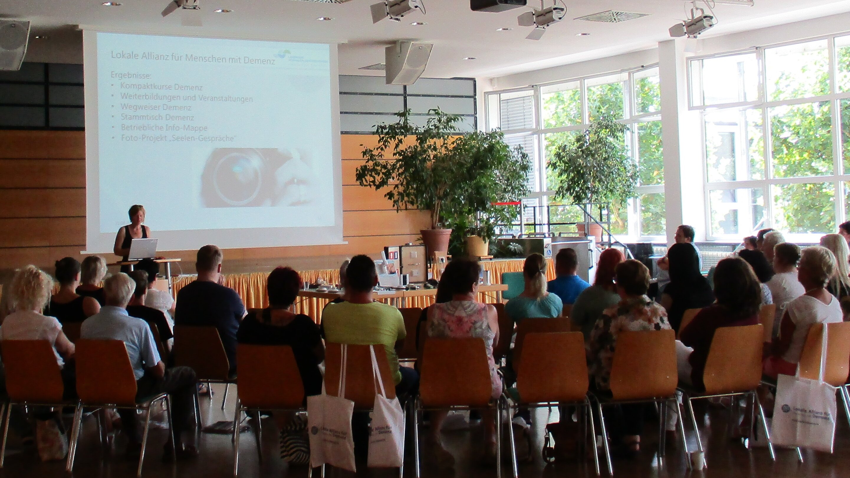 Informationsveranstaltung Foto: Großer Saal, in dem viele Menschen im Halbkreis sitzen. Vorne am Pult steht eine Rednerin.