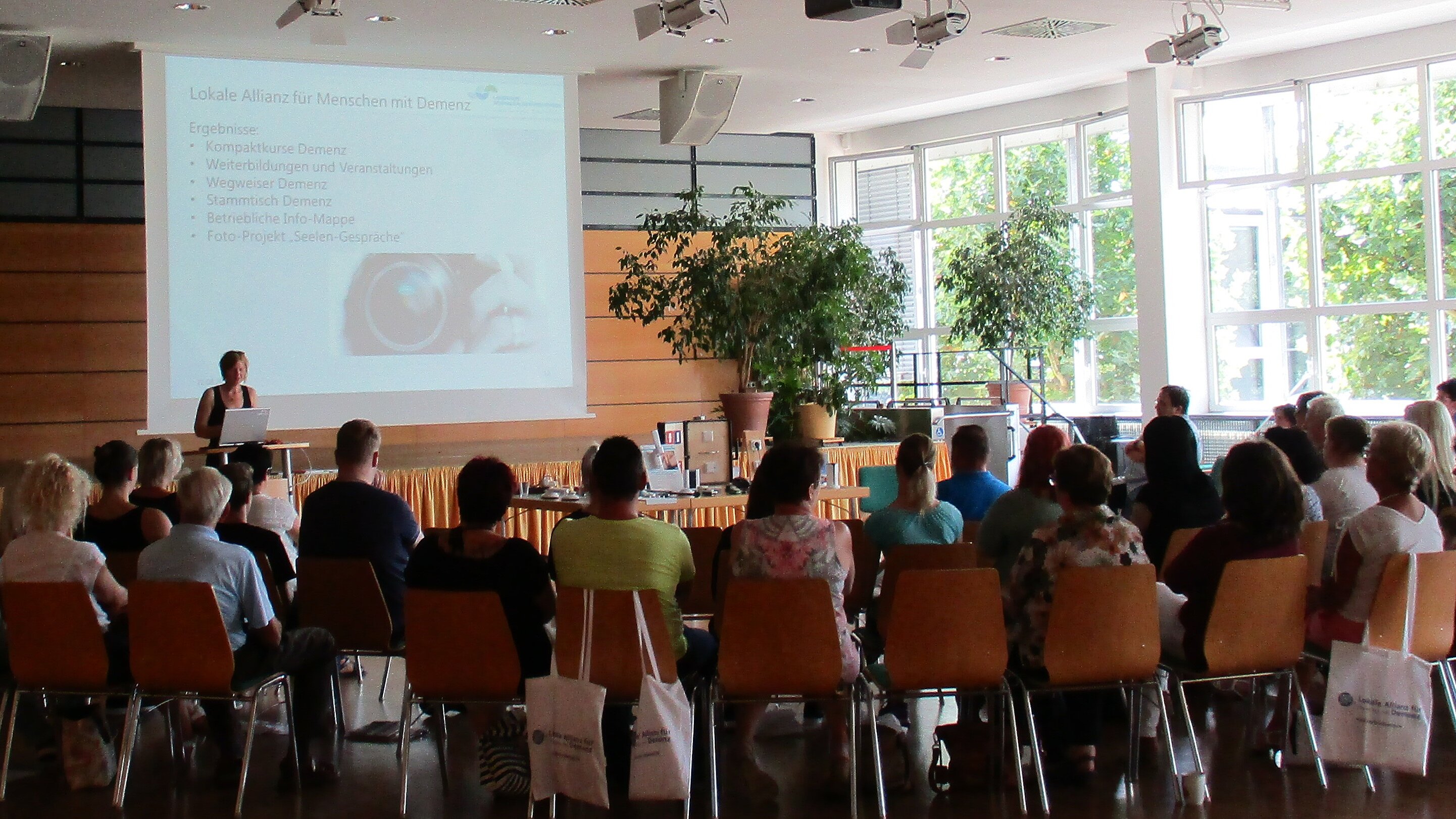 Foto: Großer Saal, in dem viele Menschen im Halbkreis sitzen. Vorne am Pult steht eine Rednerin.